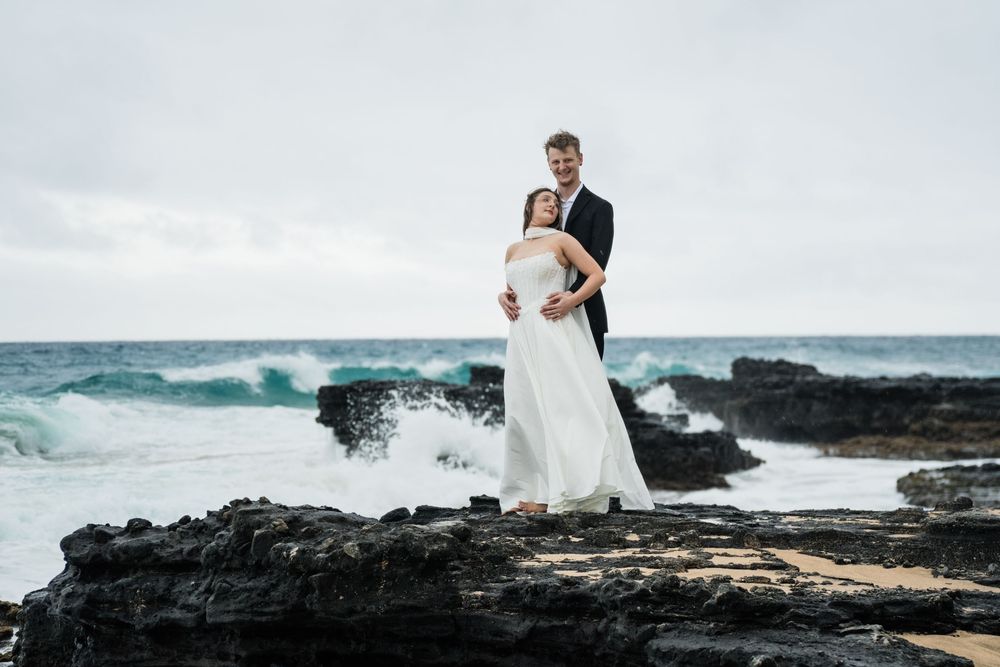 Bride and groom embrace on rocky Hawaiian coastline with dramatic ocean waves crashing behind them on a cloudy day.