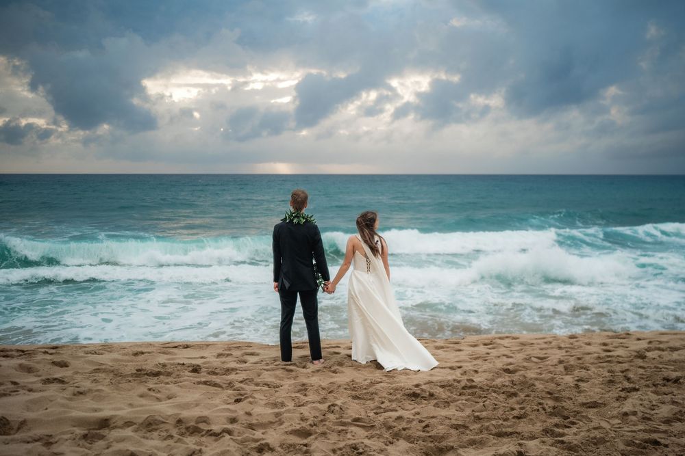 Bride and groom holding hands on a sandy beach, gazing at turquoise ocean waves under a dramatic cloudy sky.