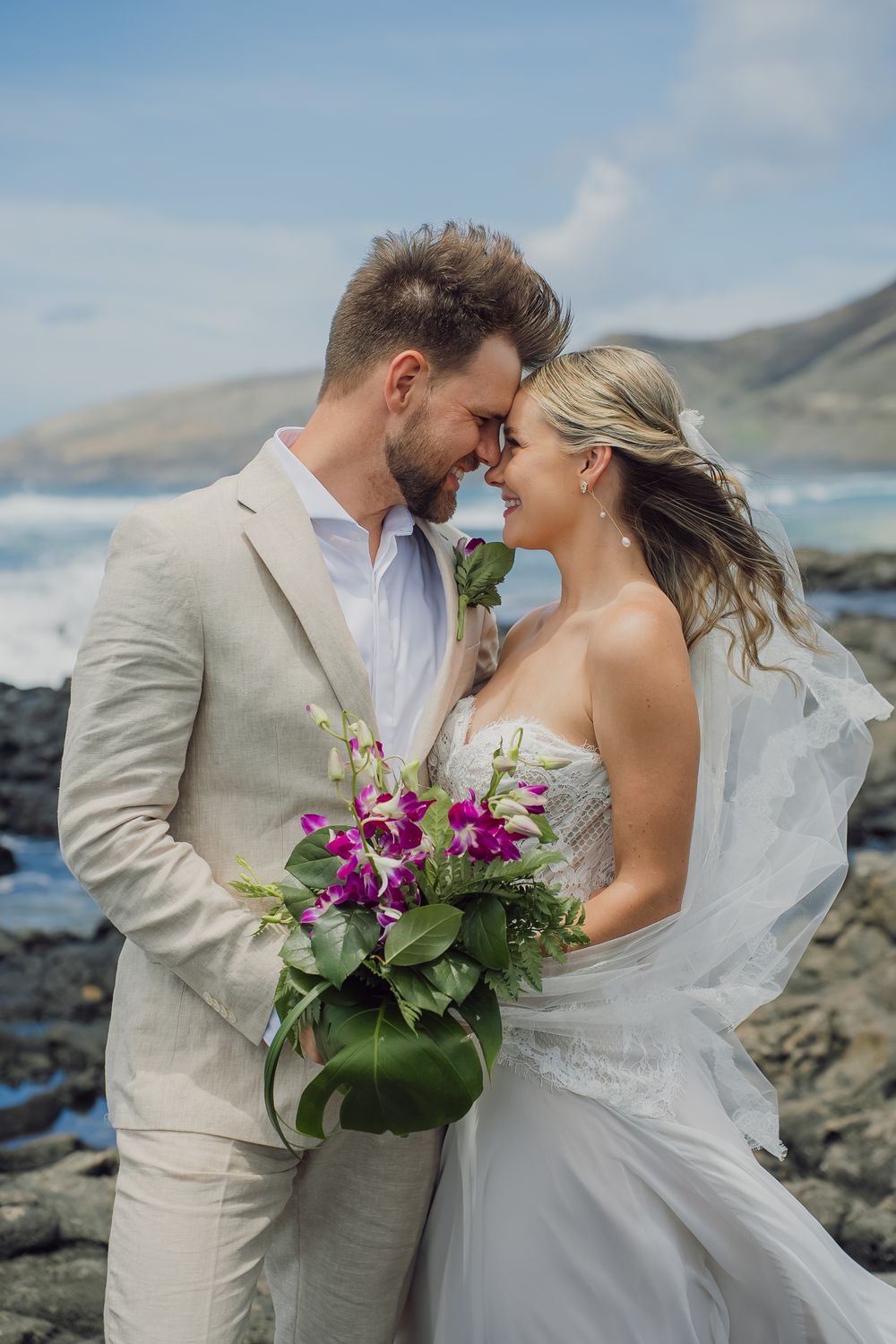 Bride and groom share a romantic moment on rocky oceanside cliffs, she holds purple tropical bouquet, wearing white gown and veil.