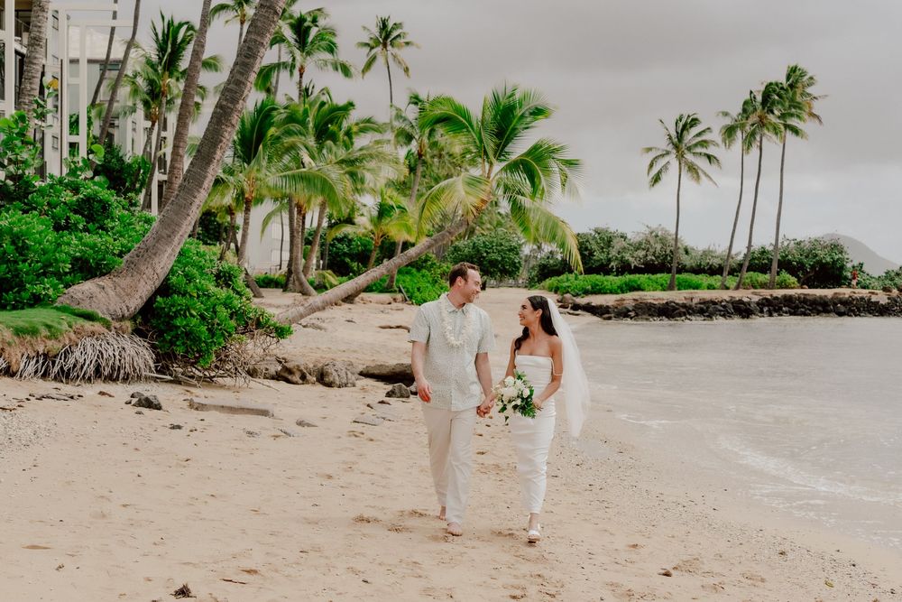 Bride and groom walk hand-in-hand on a tropical beach with palm trees, dressed in elegant white wedding attire.