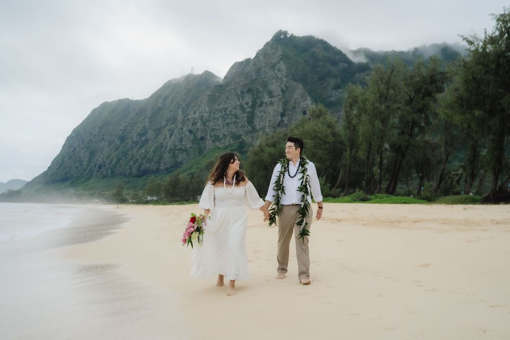 Couple in wedding attire walking hand-in-hand on a sandy Hawaiian beach with lush green mountains behind.
