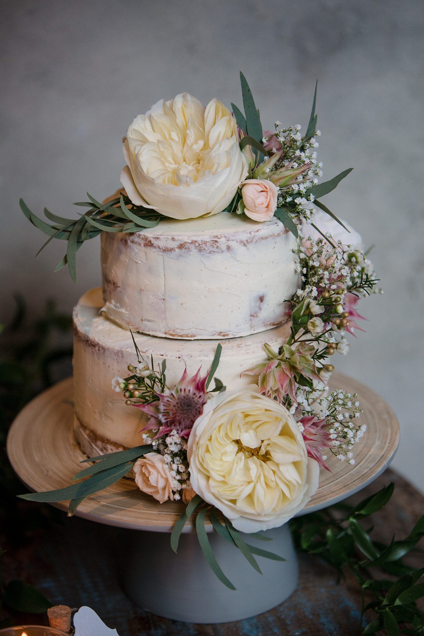 Two-tier semi-naked wedding cake adorned with white peonies, blush roses, and eucalyptus on a wooden cake stand.