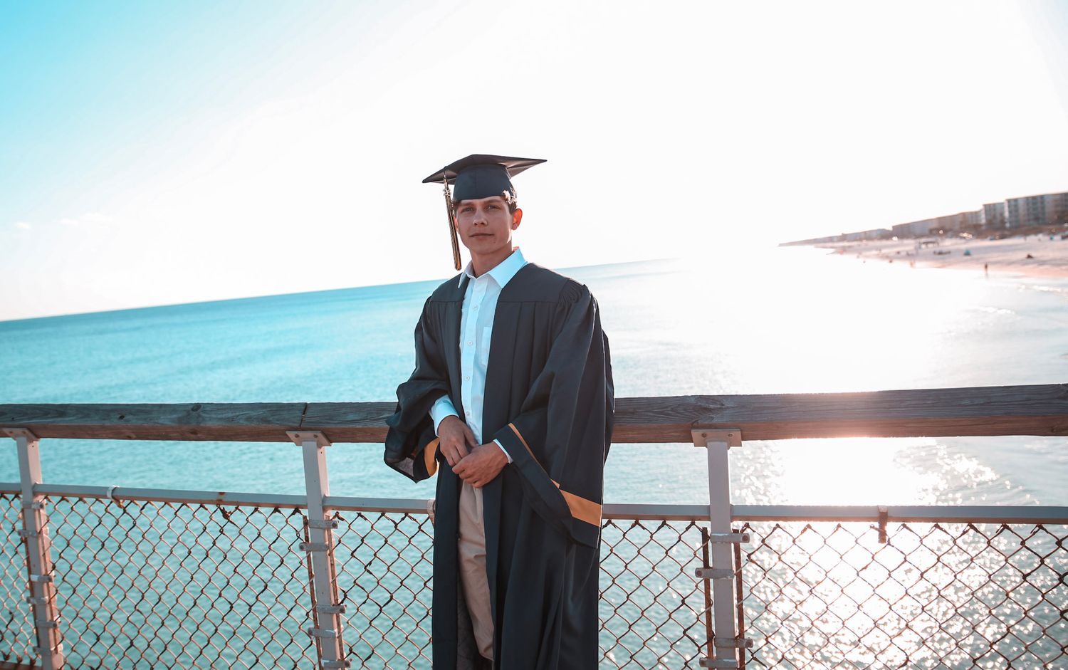 Graduate in cap and gown smiles on sunny pier overlooking bright blue ocean.