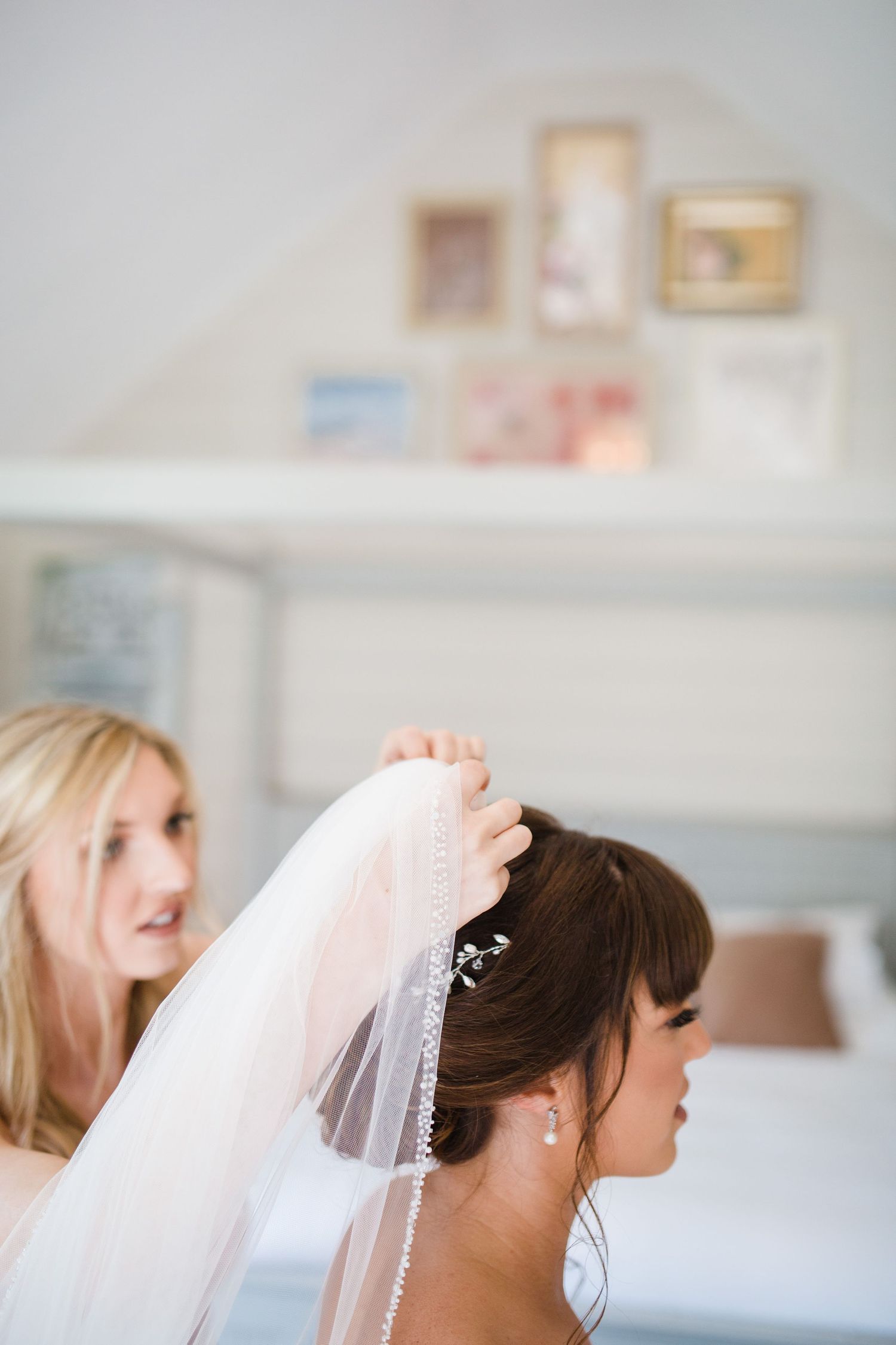 Bridesmaid placing a pearl-trimmed veil on brunette bride's updo in a bright white bedroom.