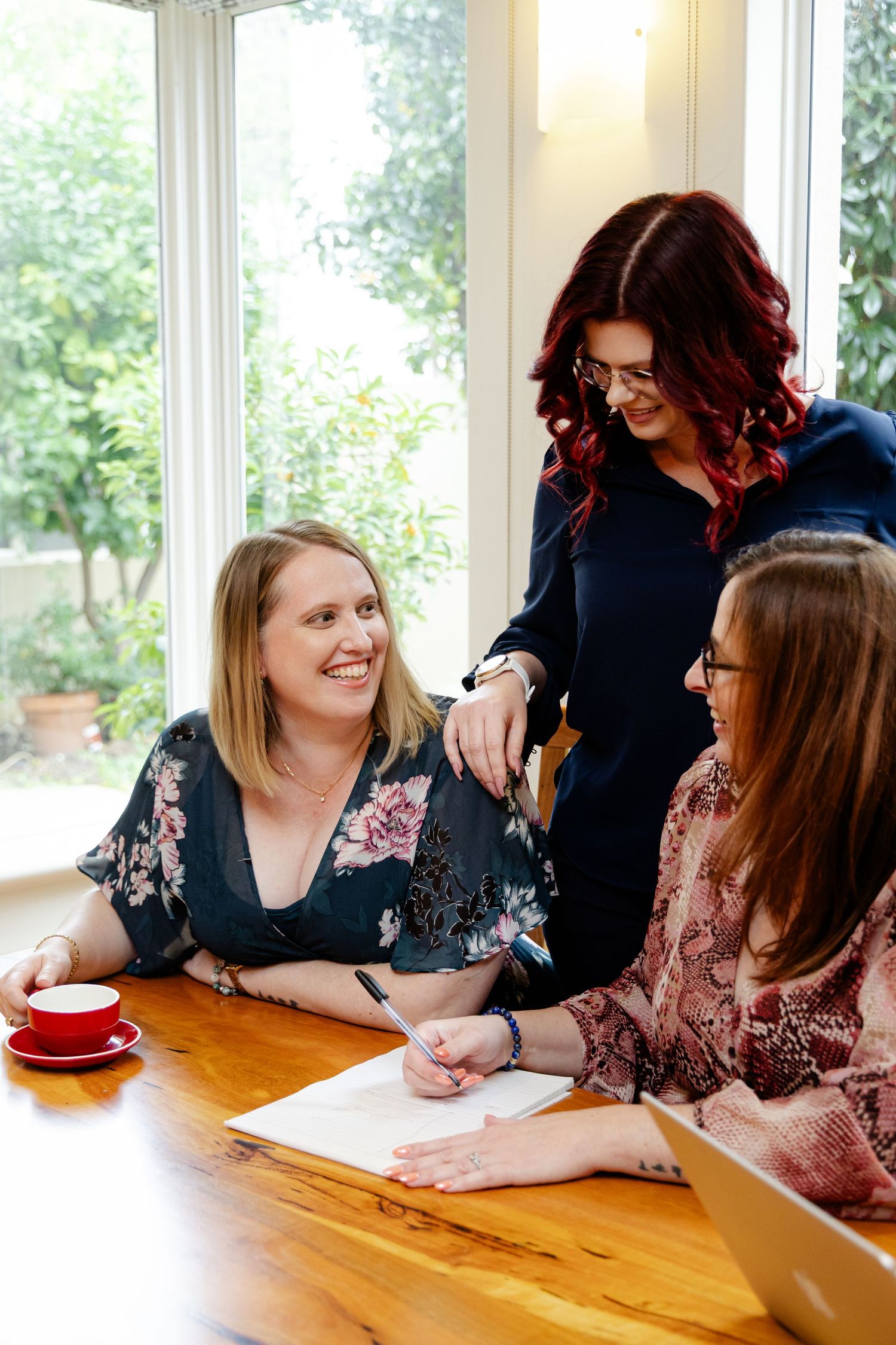 Three women collaborating at a wooden table near bright windows, smiling and reviewing documents together.