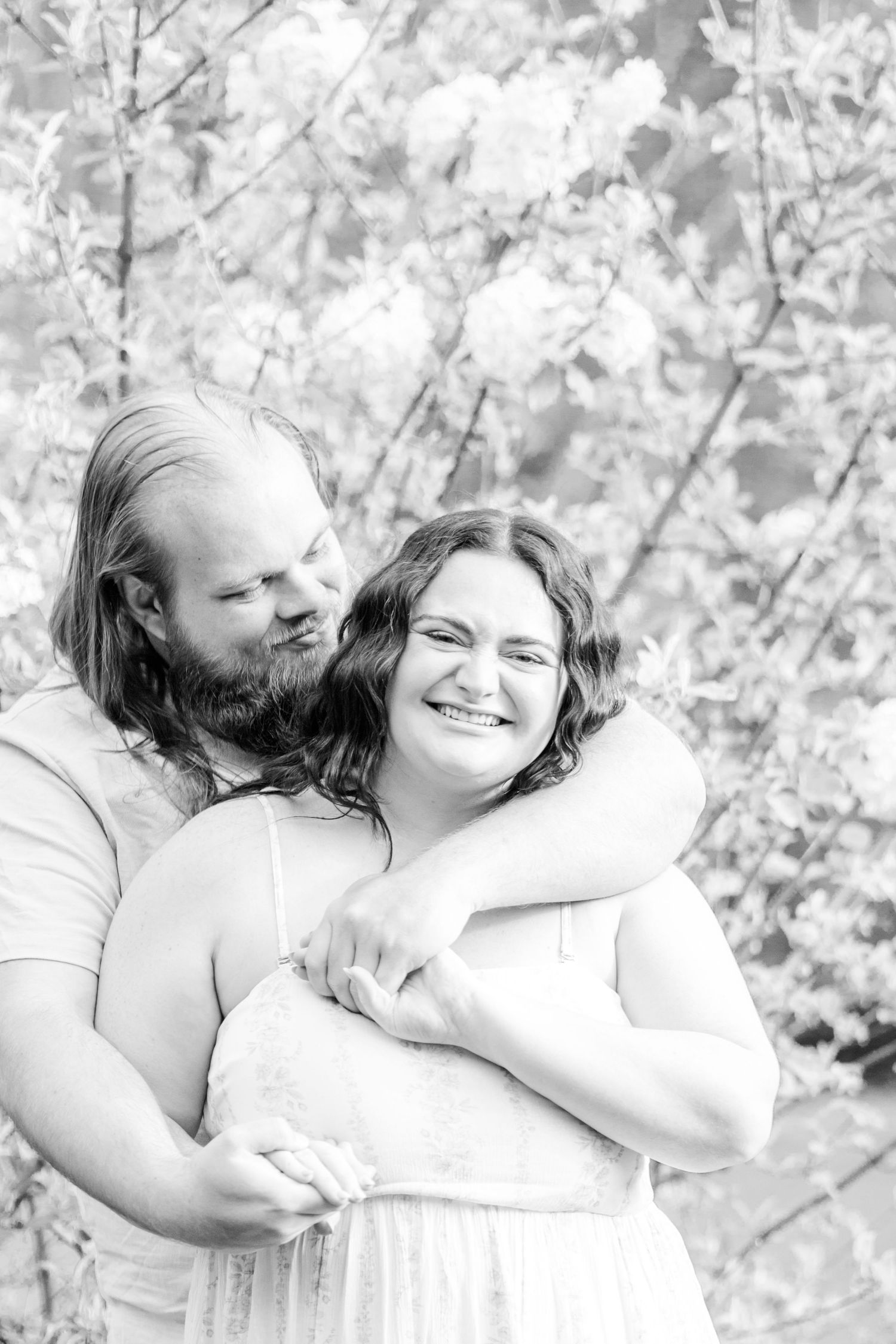 Black & white photo of a bearded man embracing, kissing a smiling woman among blooming trees. cherry blossoms wv morgantown west virginia