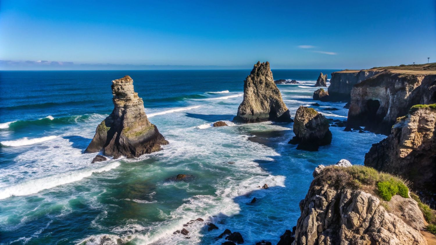 Rocky sea stacks rise from crashing ocean waves along a rugged coastline under a clear blue sky.
