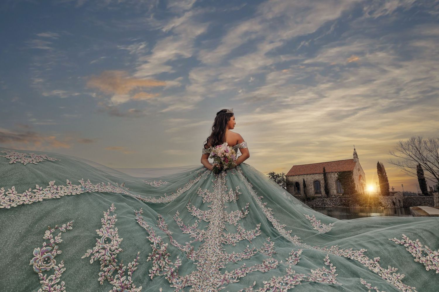 Quinceañera in a sage green gown with intricate embroidery looks toward a stone chapel at sunset, her long train fanned out behind her.