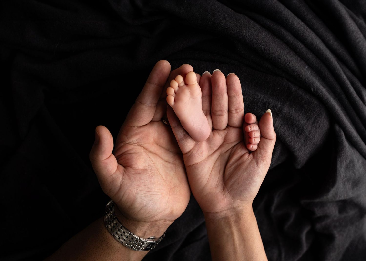 Two adult hands cradling a tiny newborn baby foot against a dark black fabric background. Elizabeth Lang Photography Surrey