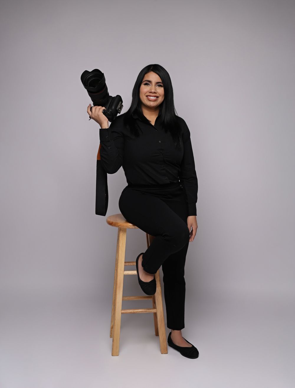 Smiling photographer in black outfit sits on stool holding camera between two studio softbox lights on white background.