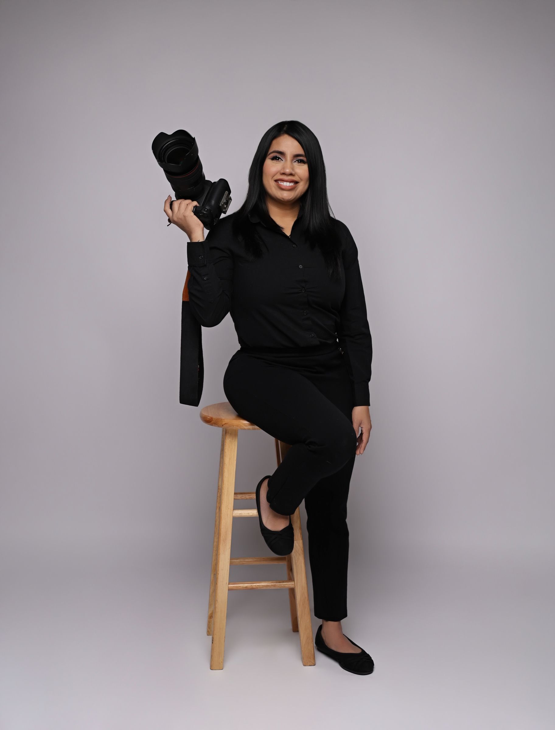 Smiling photographer in black outfit sits on stool holding camera between two studio softbox lights on white background.