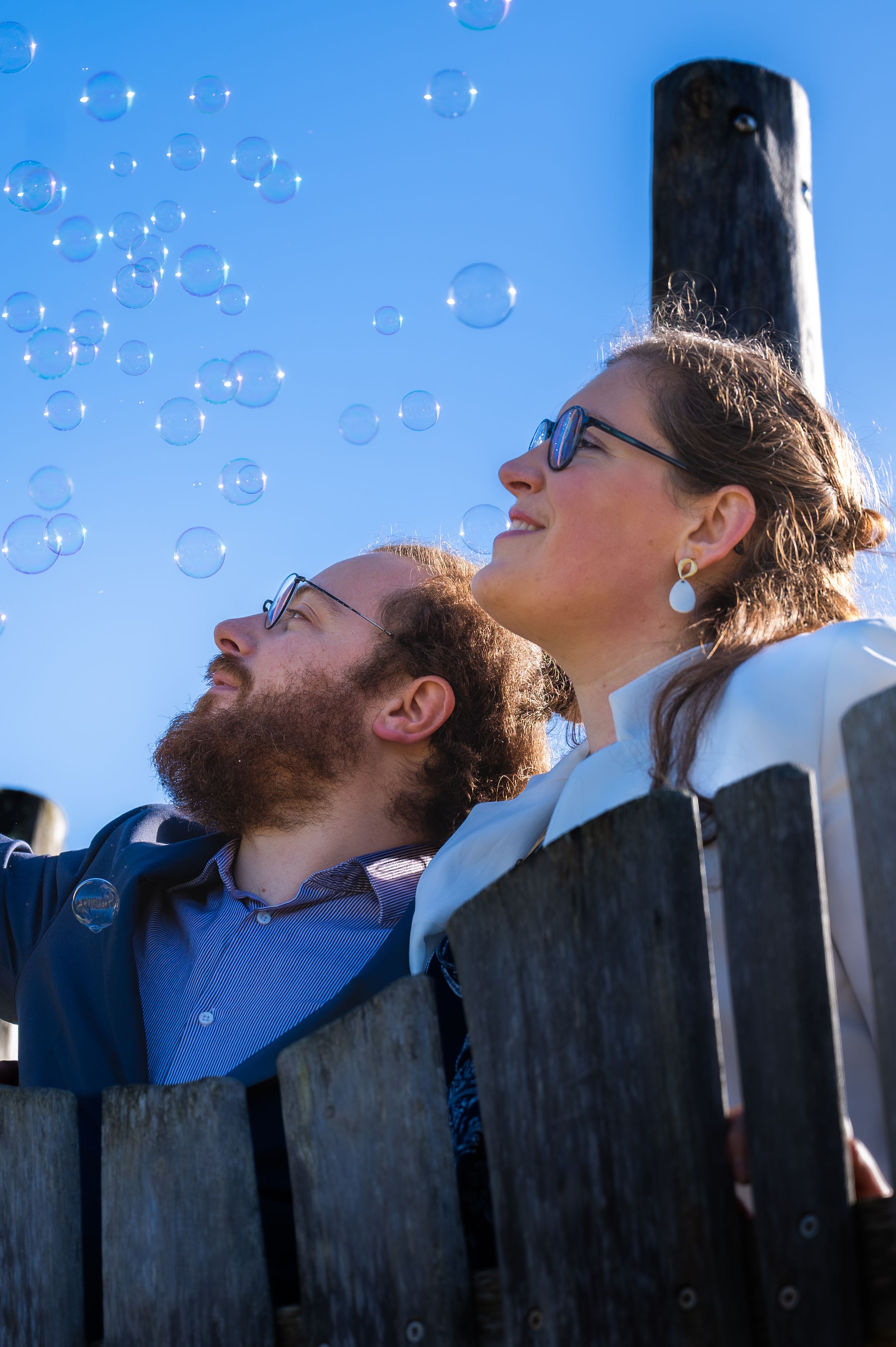 Un couple souriant, levant les yeux vers des bulles de savon qui flottent dans un ciel d'un bleu éclatant, près d'une clôture en bois.