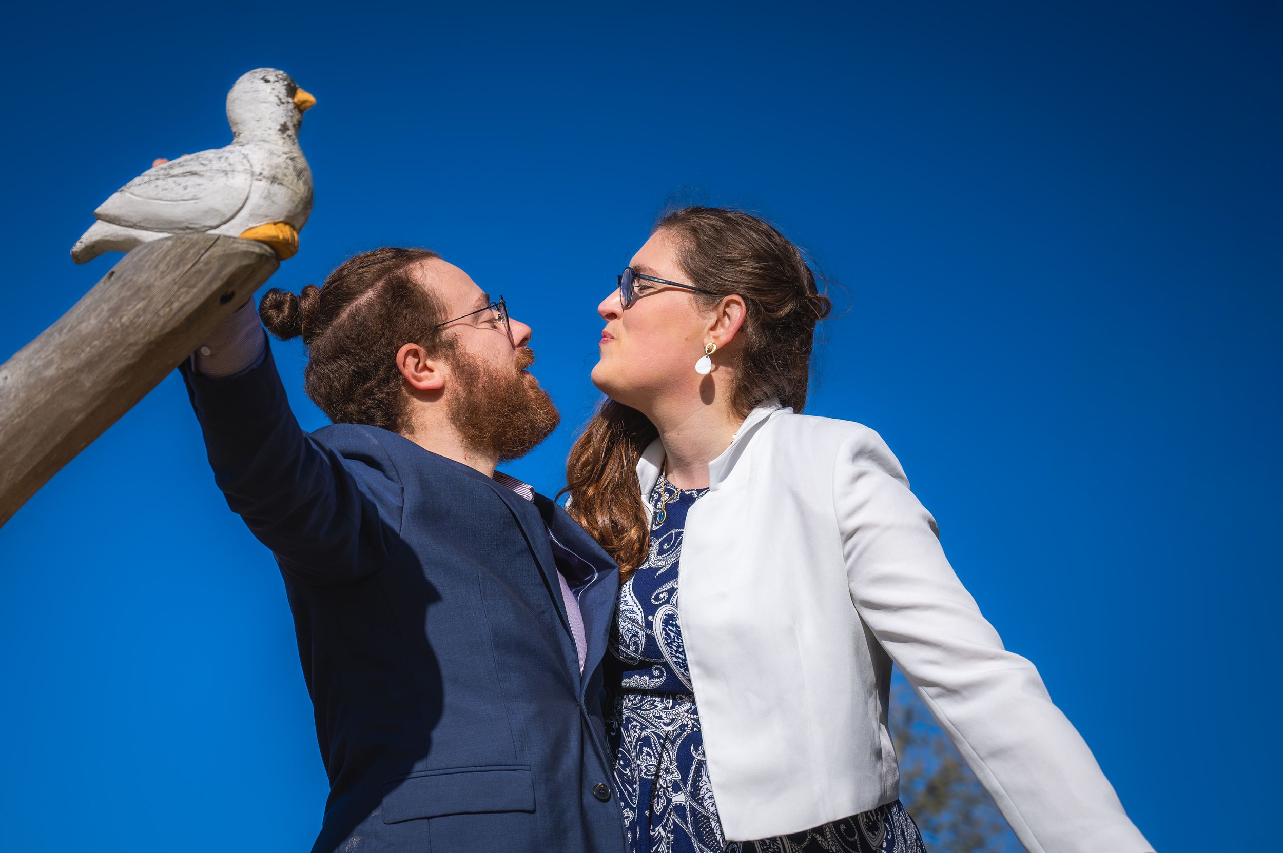 Un couple heureux se regardant, avec un oiseau blanc perché sur un poteau, sur fond de ciel bleu.