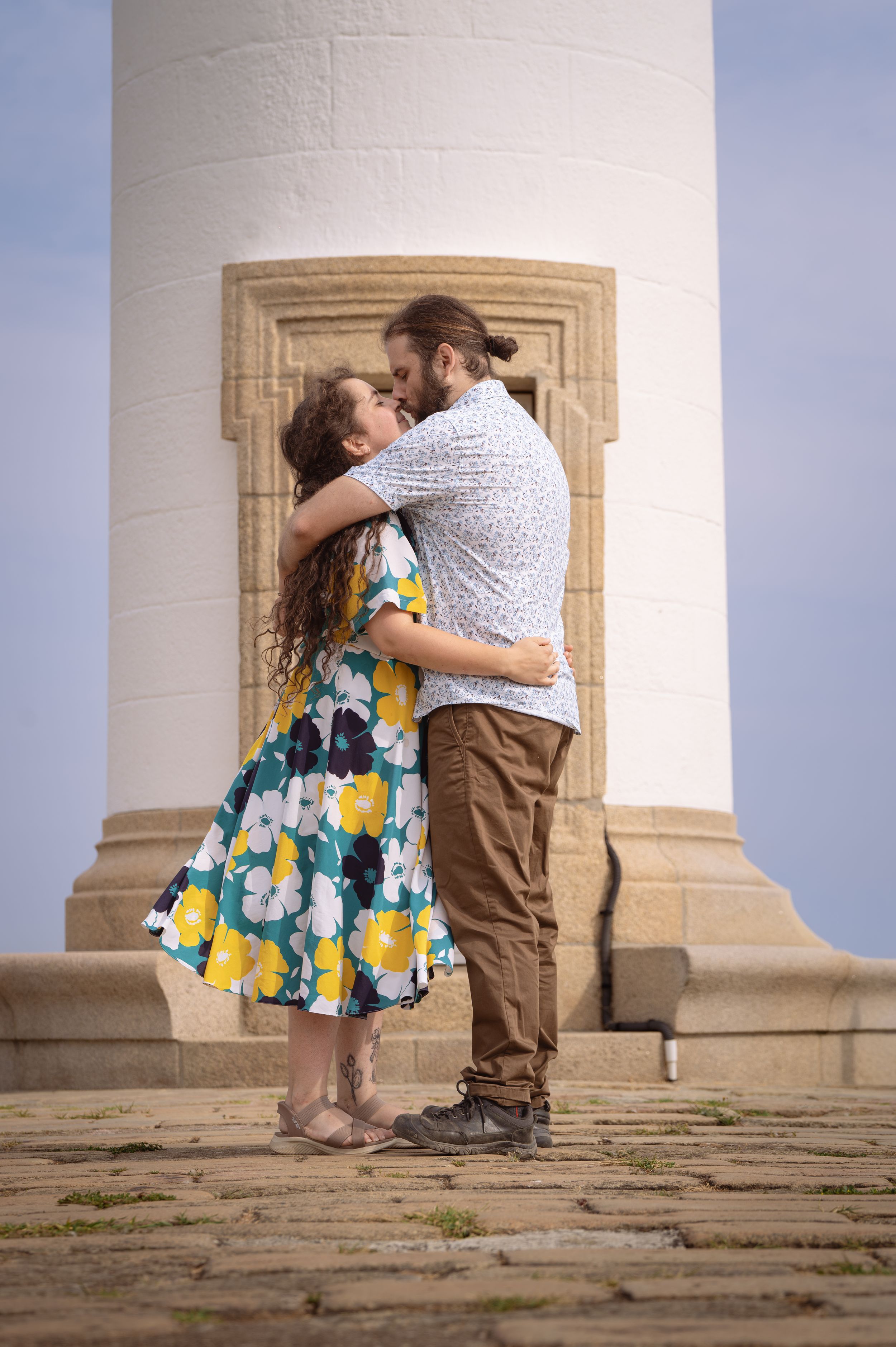 Un couple en robe fleurie et chemise décontractée s'embrassant devant un phare blanc par une journée ensoleillée.