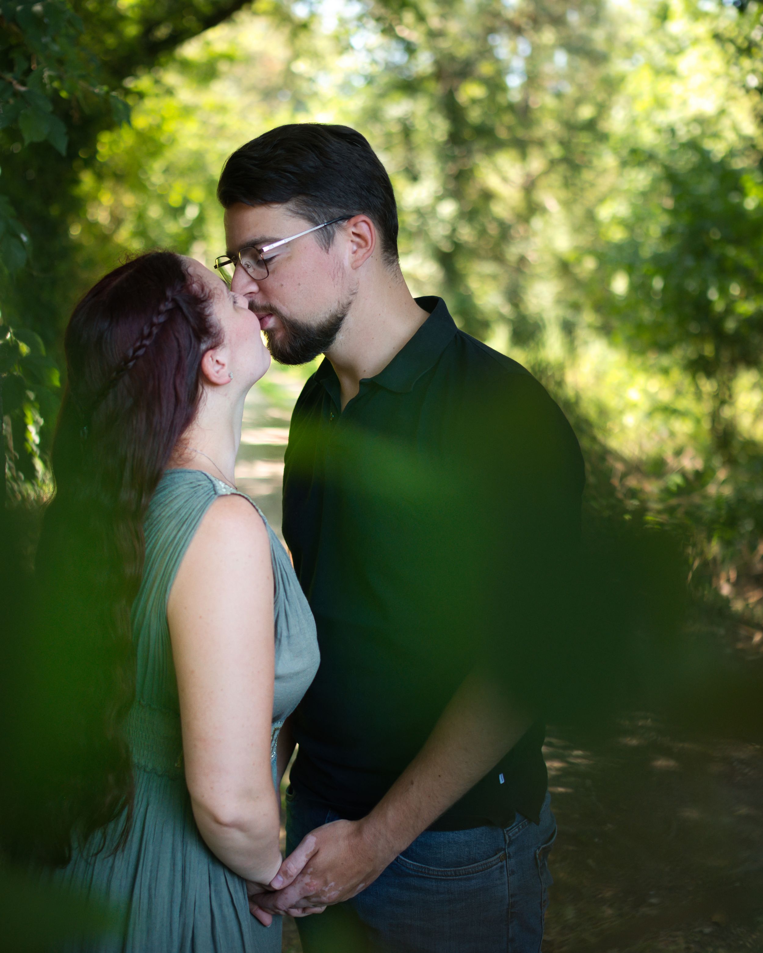 Couple sharing a romantic kiss while holding hands on a sunlit forest path.