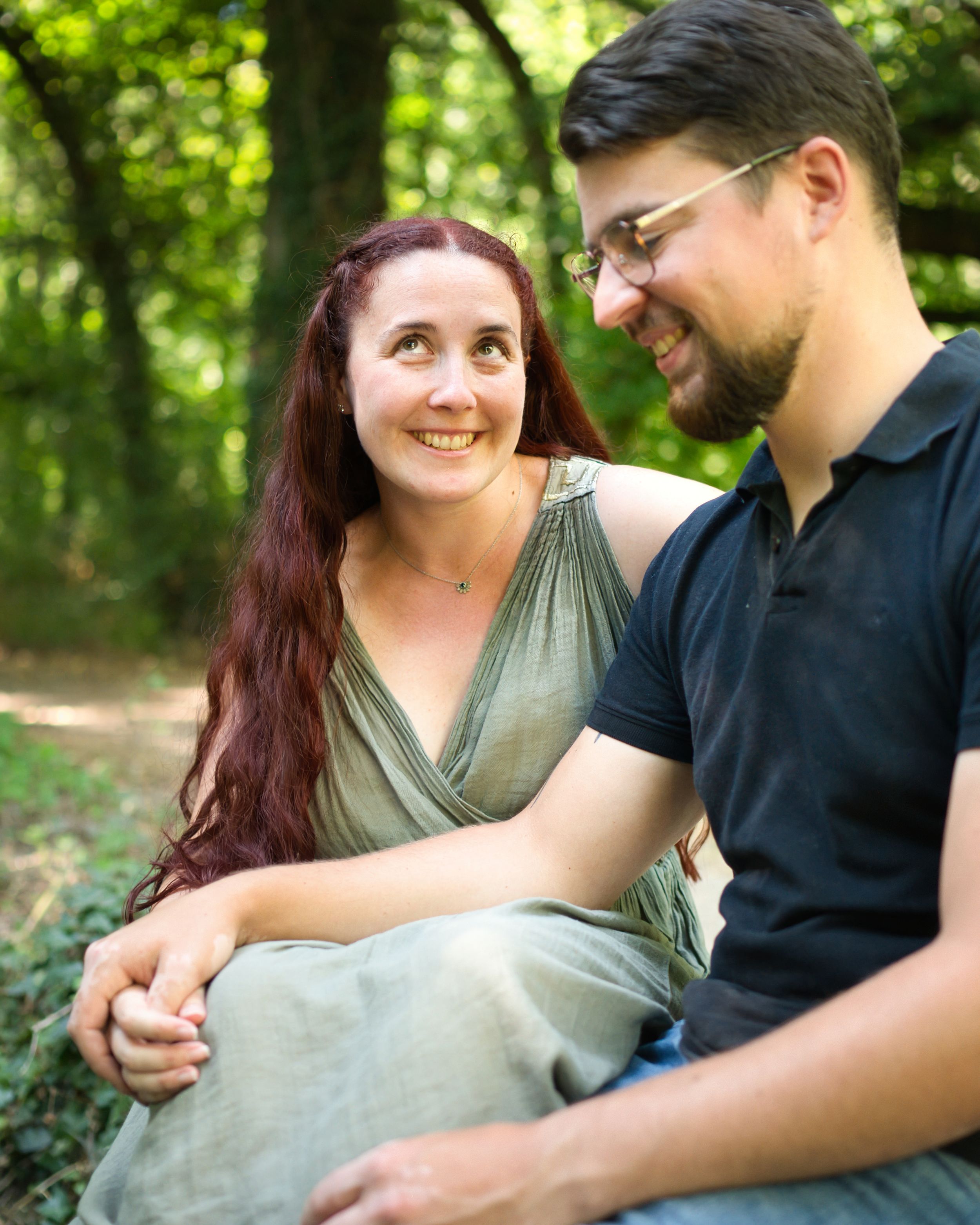 Un couple souriant, assis côte à côte dans une forêt verdoyante, se tenant la main et se regardant avec tendresse.