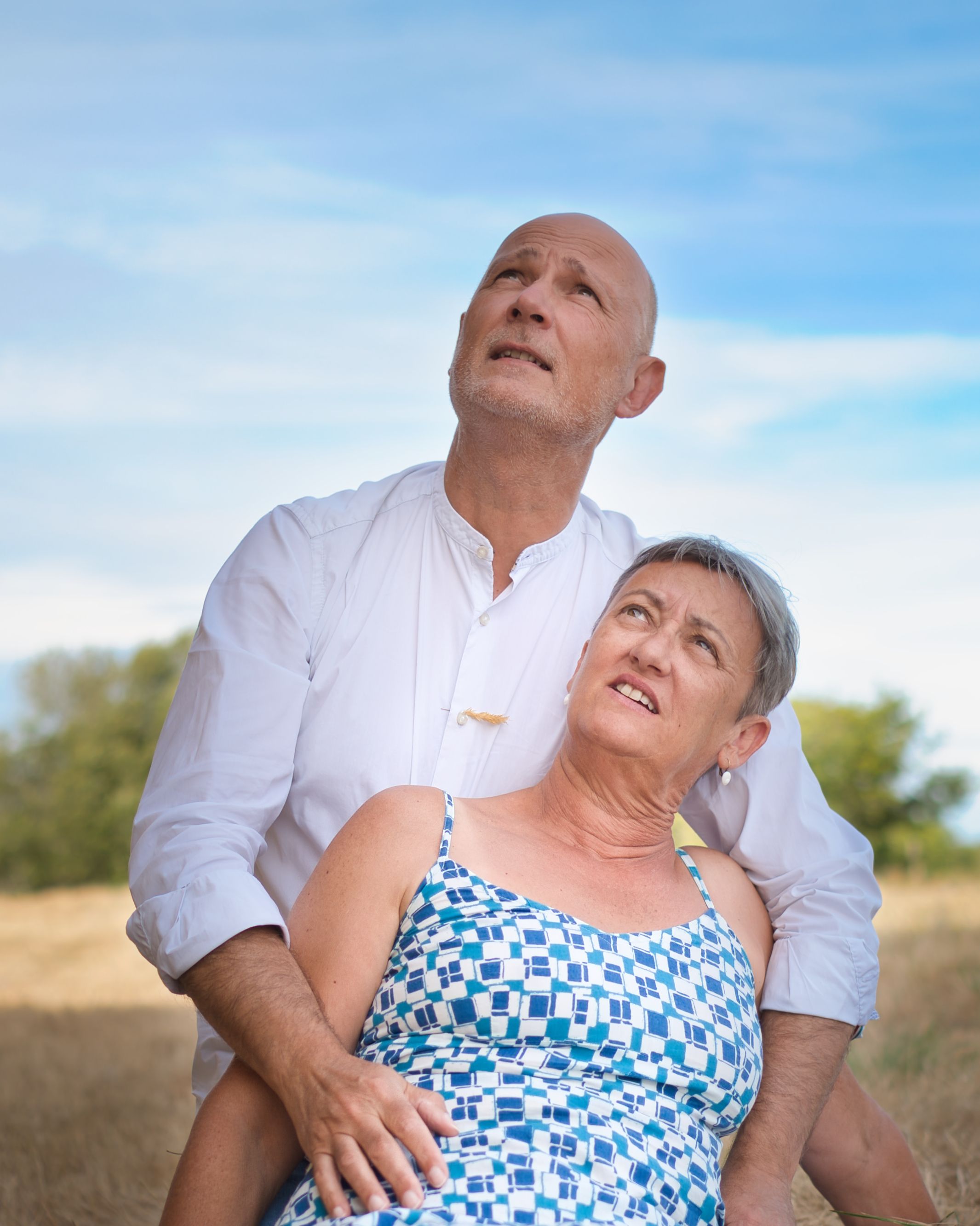 Un couple âgé en plein air, dans un champ doré, le regard tourné vers le ciel bleu parsemé de nuages blancs.