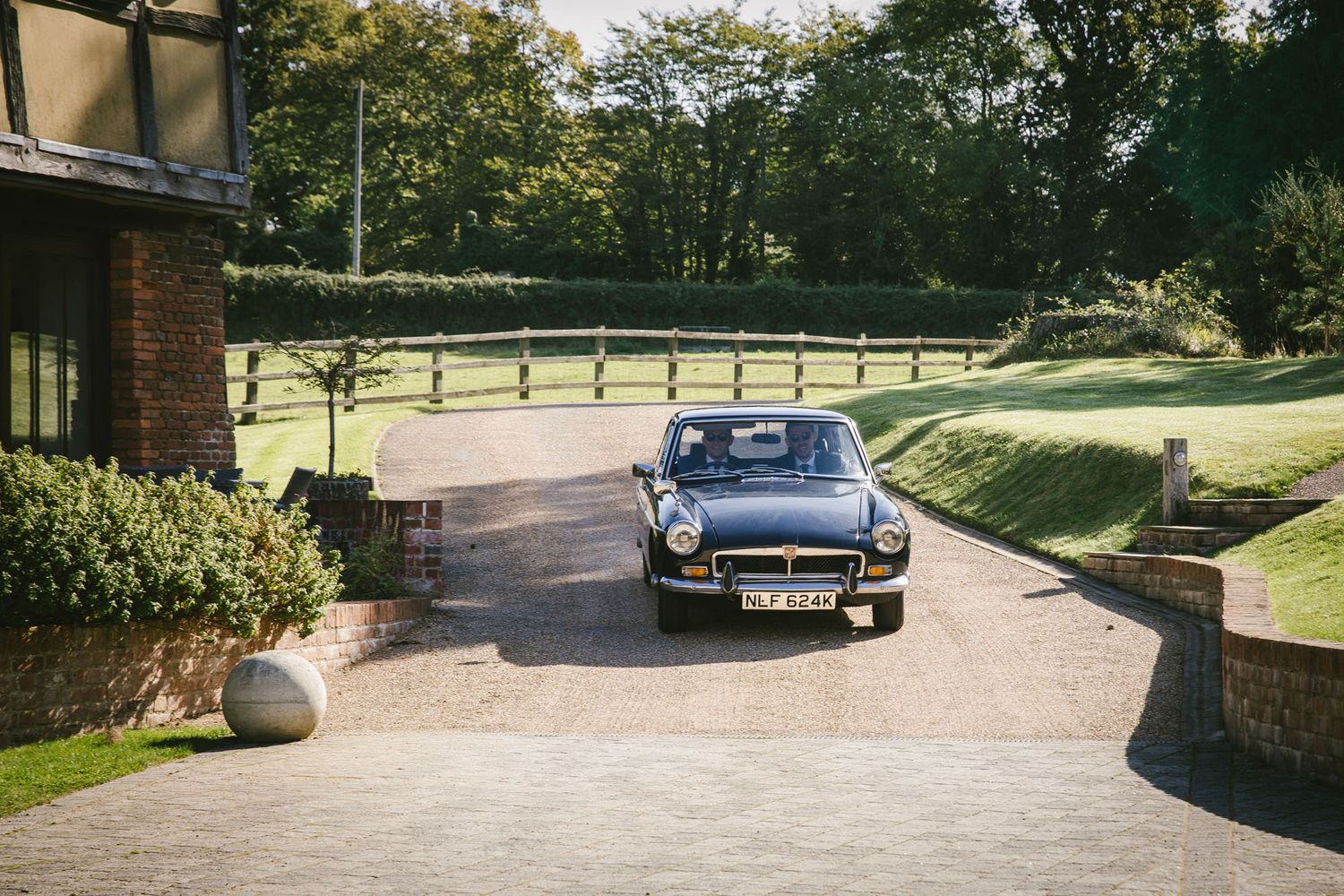 A classic black MGB sports car parked on a gravel driveway surrounded by lush greenery and wooden fencing.