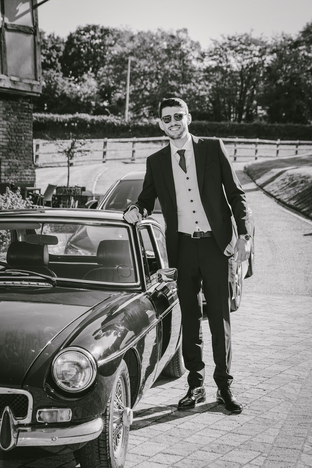 Black and white photo of a well-dressed man in a suit and sunglasses posing beside a classic vintage convertible car.