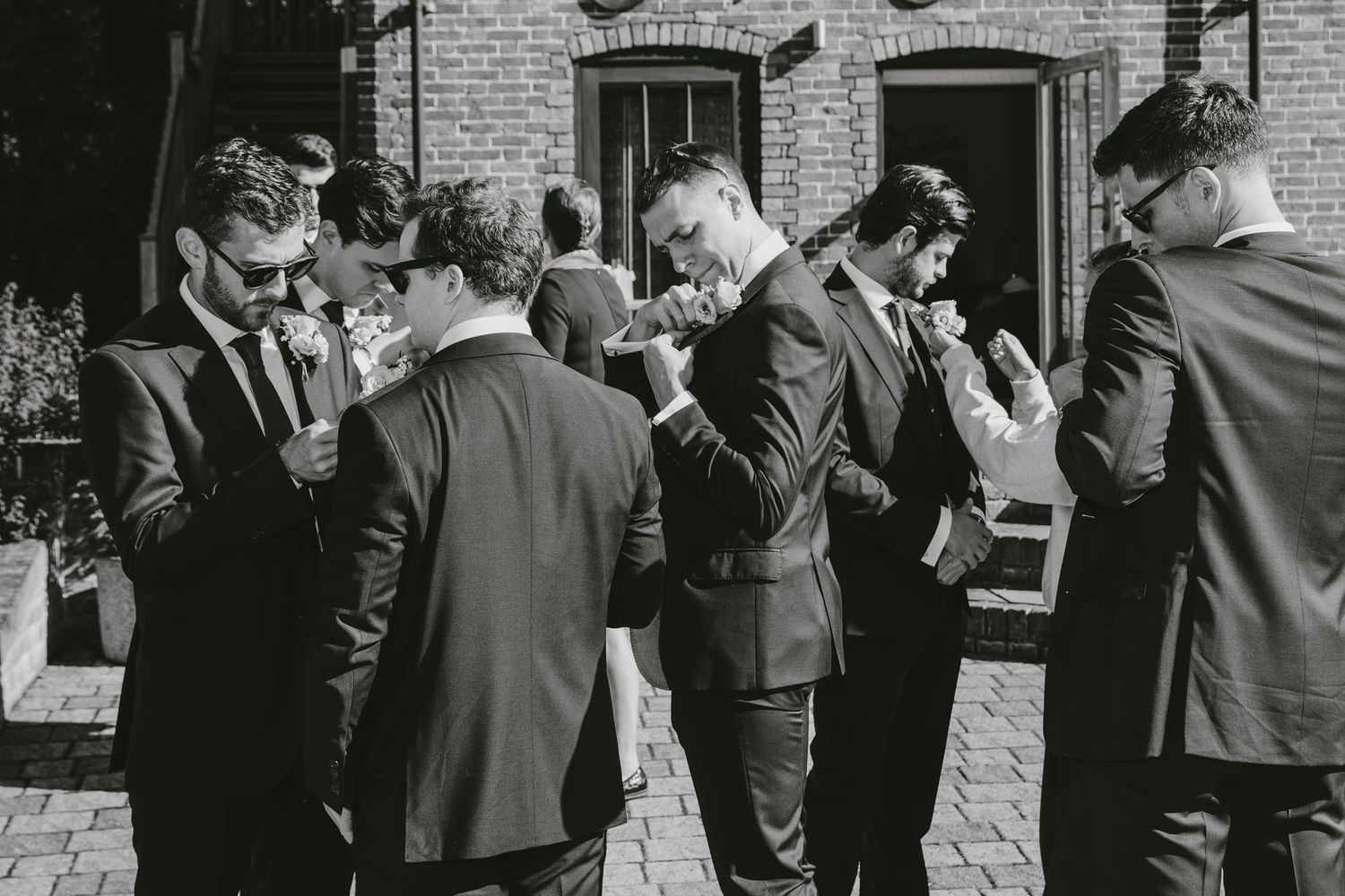Groomsmen in dark suits adjusting boutonnieres outdoors at a wedding venue in black and white photo.