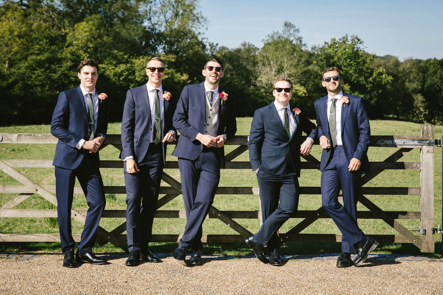 Five groomsmen in navy suits and sunglasses pose by a wooden fence gate outdoors on a sunny wedding day.