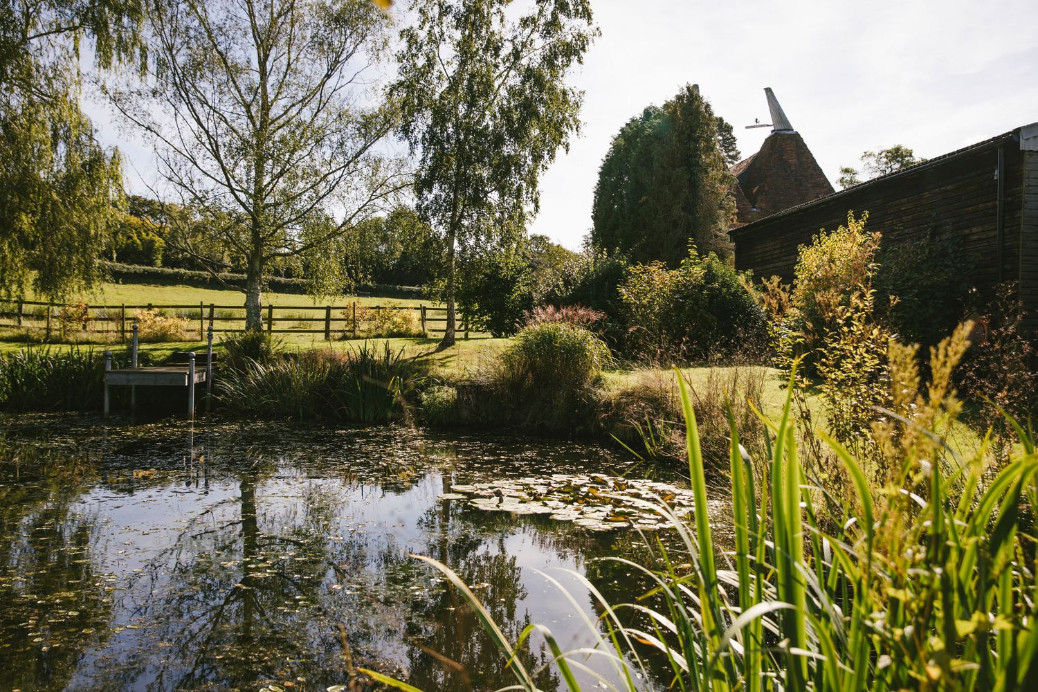 Tranquil garden pond with lily pads, lush greenery, willow trees, and a rustic brick oast house on a sunny day.