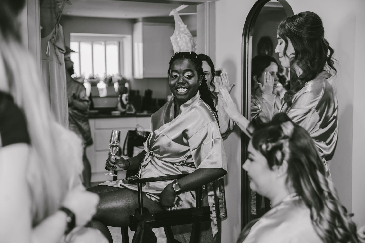 Bridesmaids laughing and getting ready together, one wearing a face mask and satin robe, in black and white.