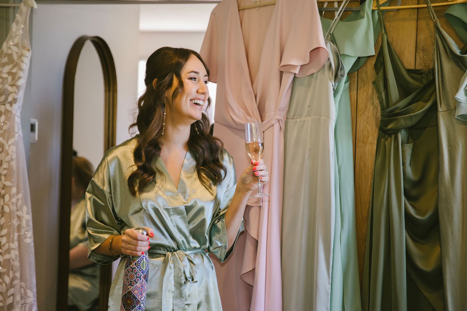Smiling bride in sage green robe holding champagne glass surrounded by hanging bridesmaid dresses in soft pink and green.