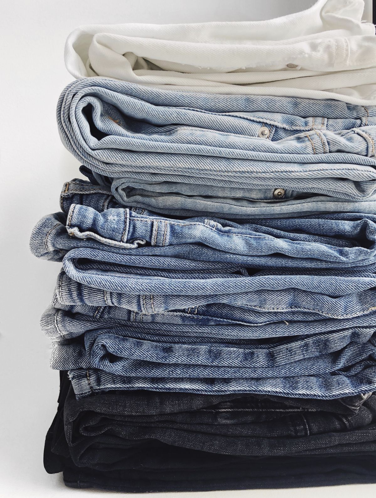 Stack of folded jeans in various shades from white to dark navy, neatly piled on a white surface.
