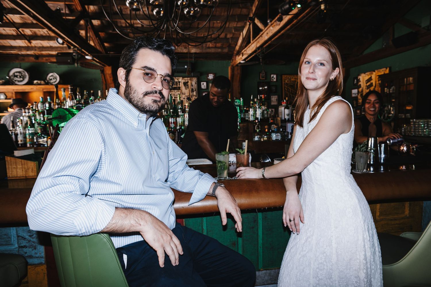 A man and woman pose together at a stylish bar, with shelves of bottles and a bartender visible in the background.