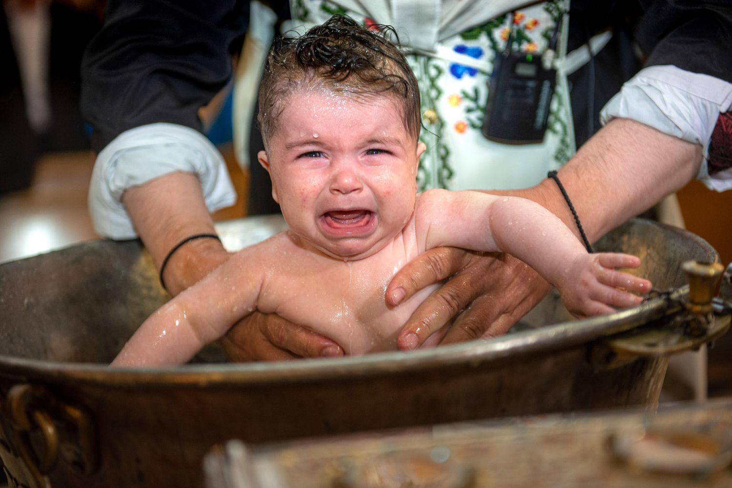 Crying baby being baptized in a traditional bronze baptismal font during christening.