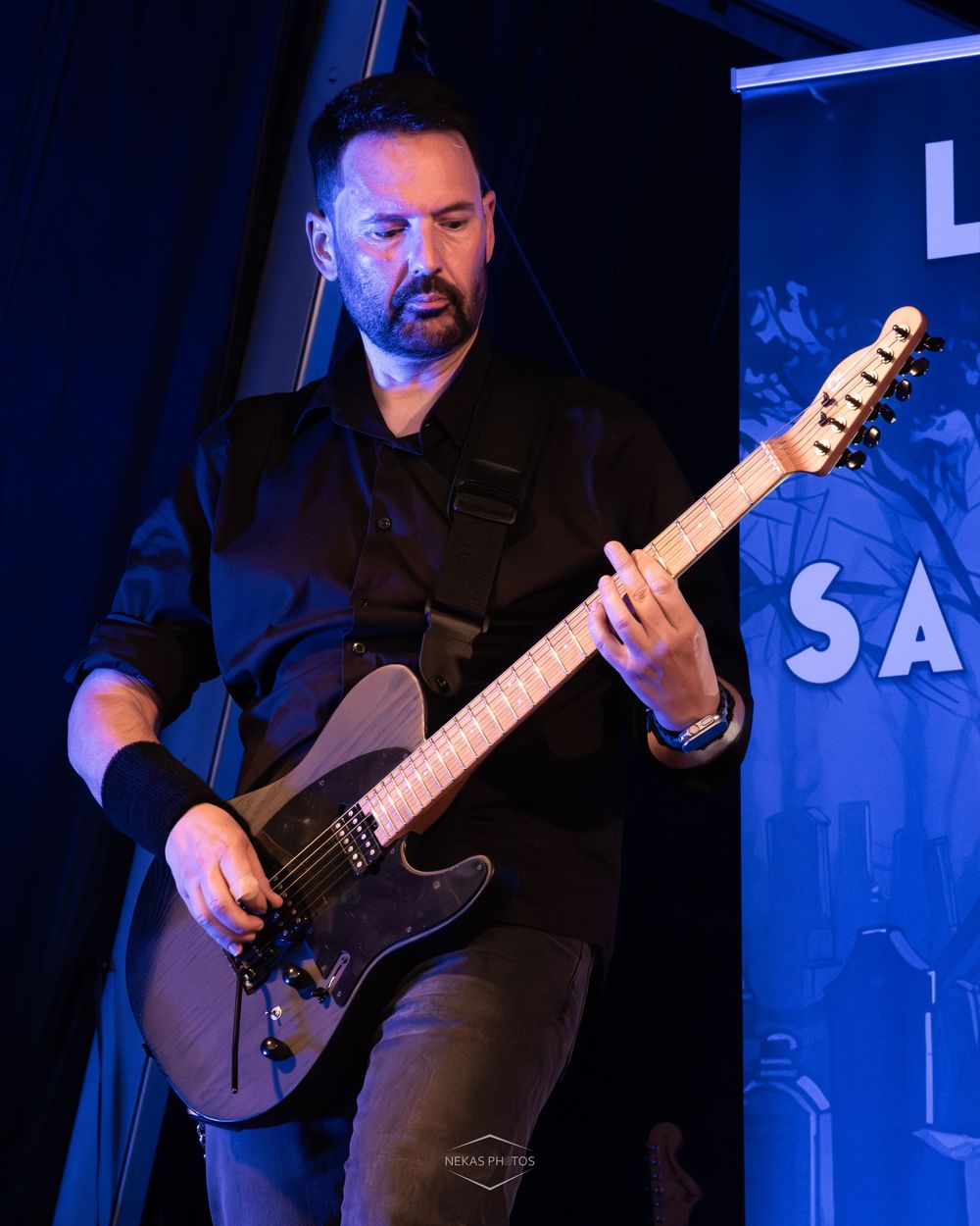 Musician in black shirt playing electric guitar on stage under blue lighting at a live performance.