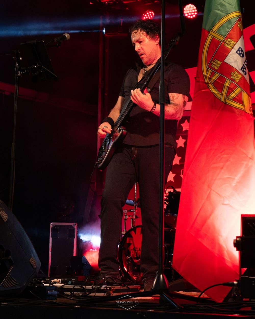Guitarist performing on stage in black outfit under red lights with Portuguese flag backdrop.