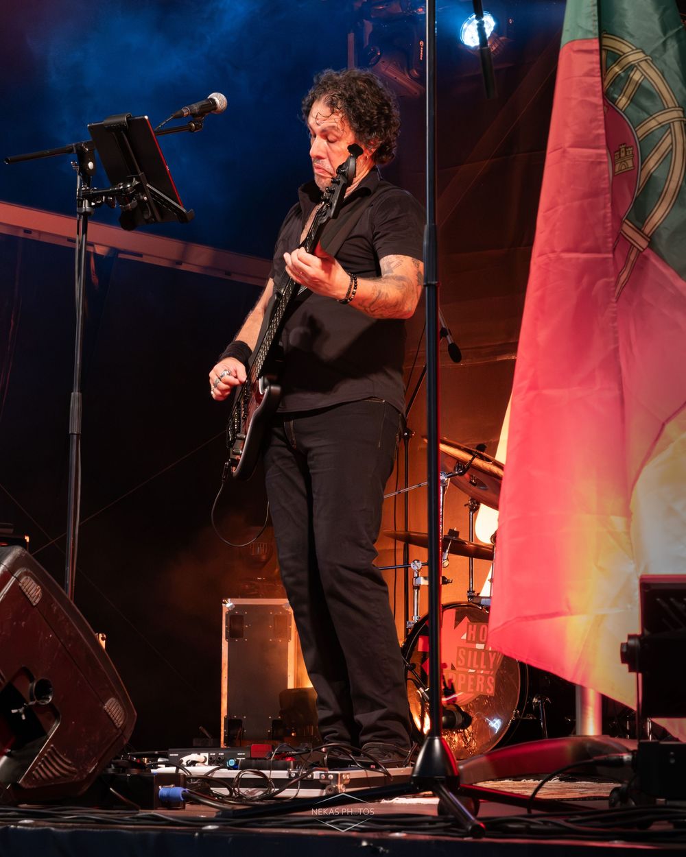A musician in all-black attire plays electric guitar on a dimly lit stage with colorful lighting and a music stand nearby.