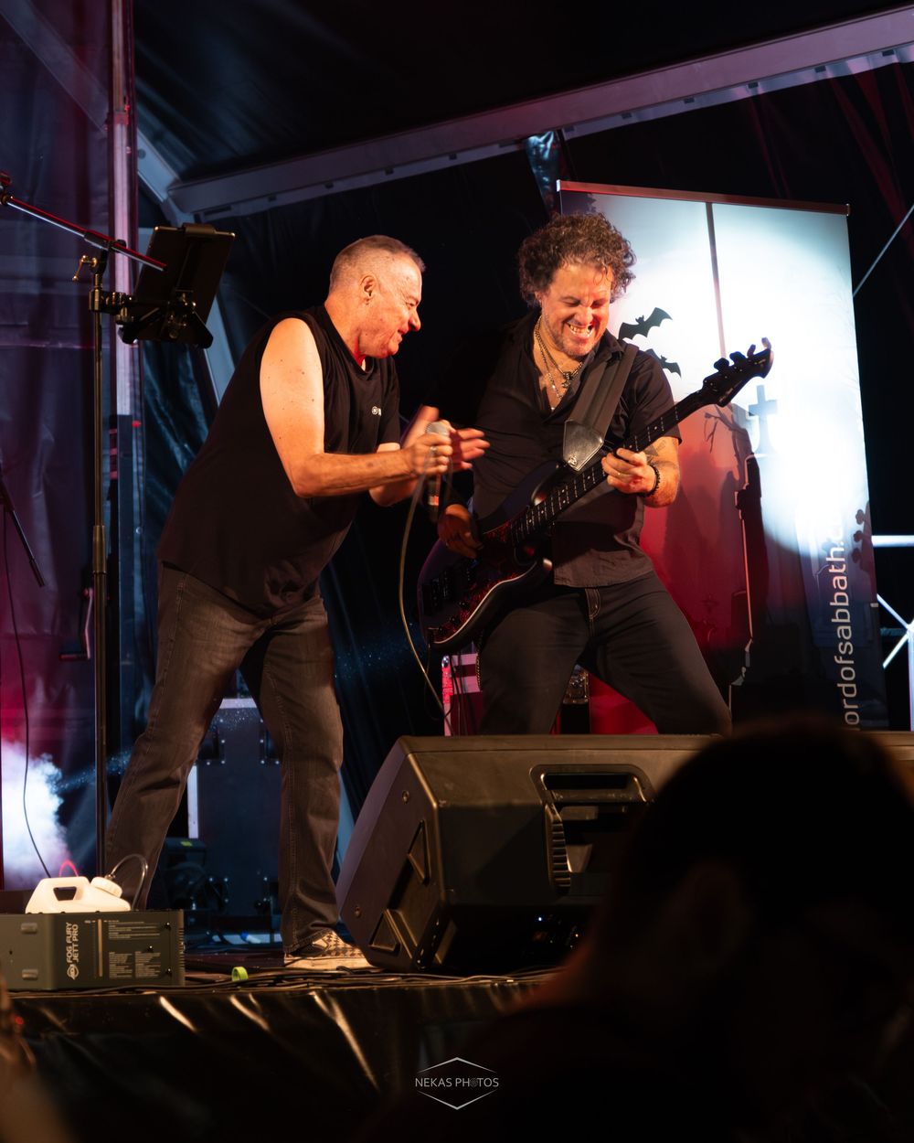 Two musicians perform energetically on stage, one singing and one playing electric guitar, both dressed in black.