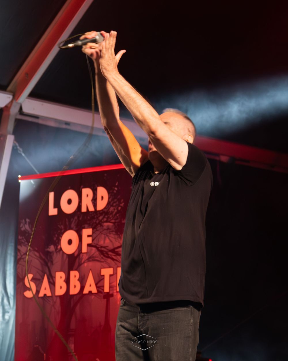 A bald vocalist performs on stage holding a microphone aloft before a Lord of Sabbath banner under red stage lighting.