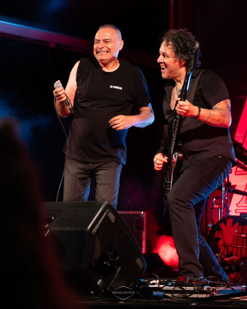 Two musicians perform on stage, one singing into a mic and one playing guitar, both wearing black shirts under red lights.