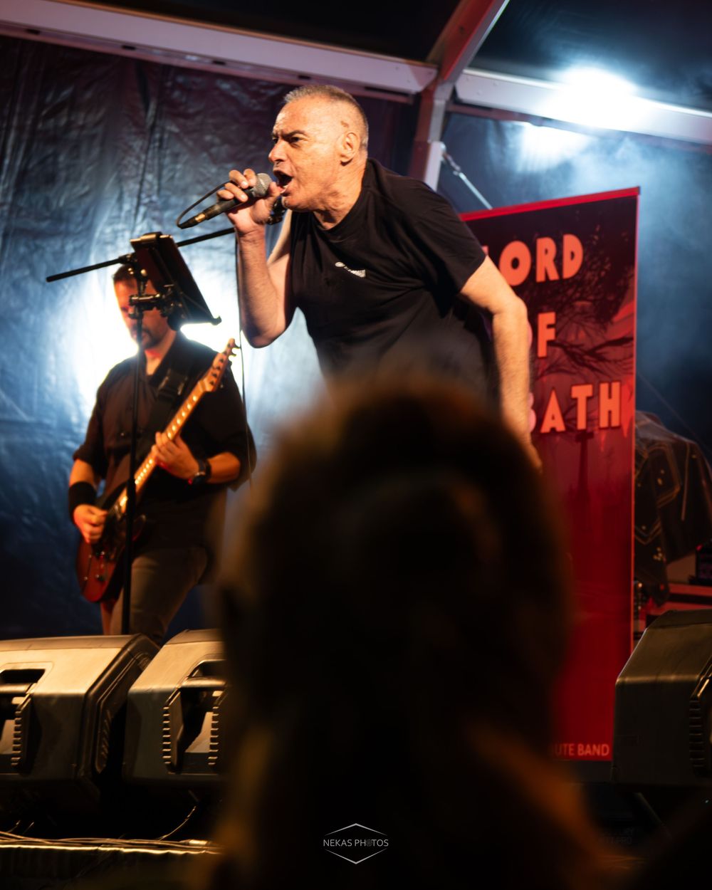A bald vocalist performs intensely on stage with a microphone while a guitarist plays behind him at a live rock concert.