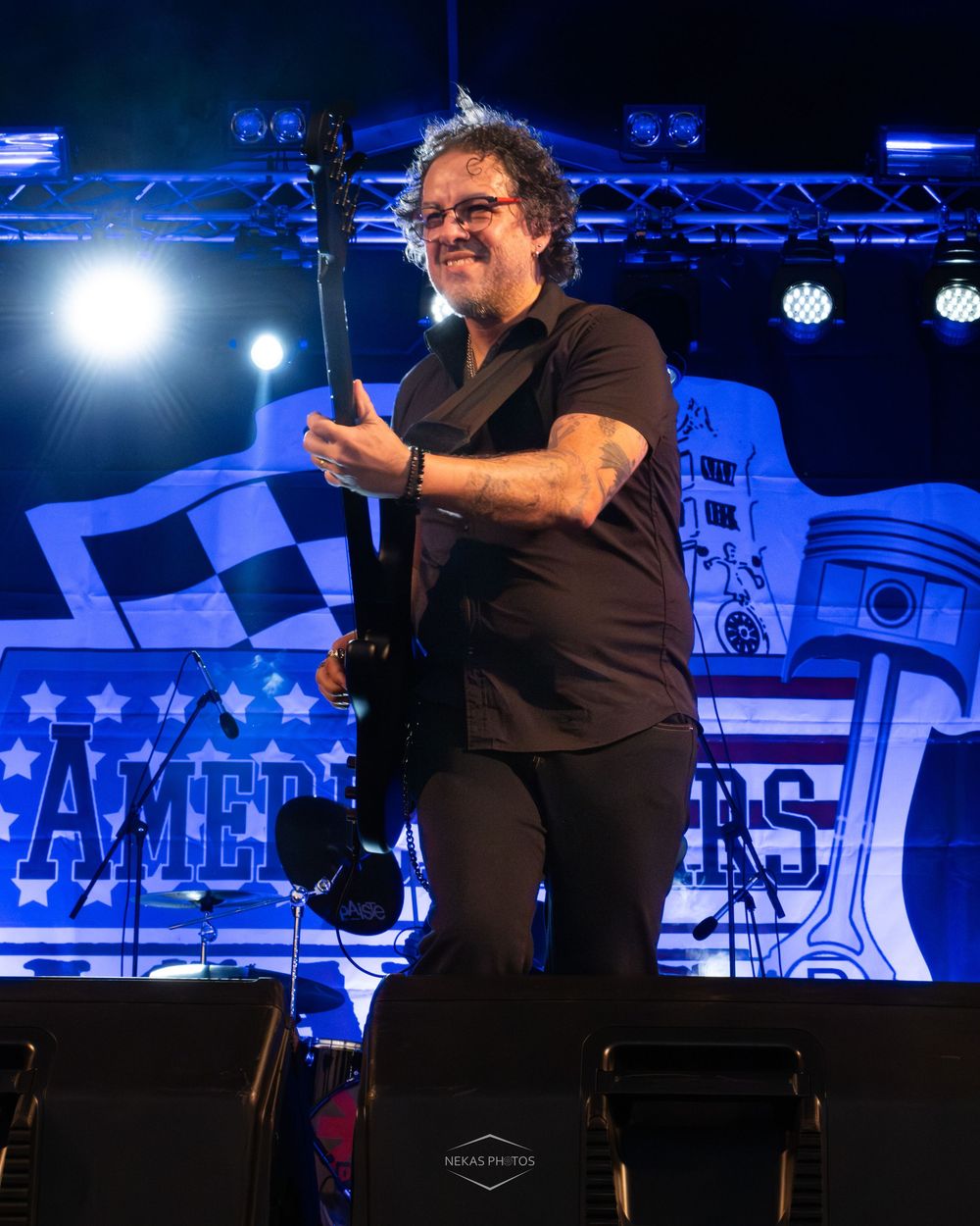 A smiling guitarist in black performs on stage with an American-themed blue backdrop at an outdoor concert.