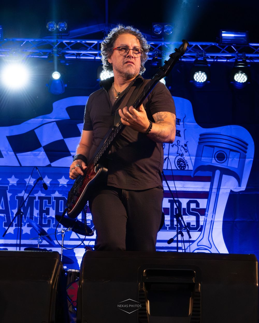 Male guitarist performing on stage with electric guitar at outdoor rock concert with blue stage lighting.