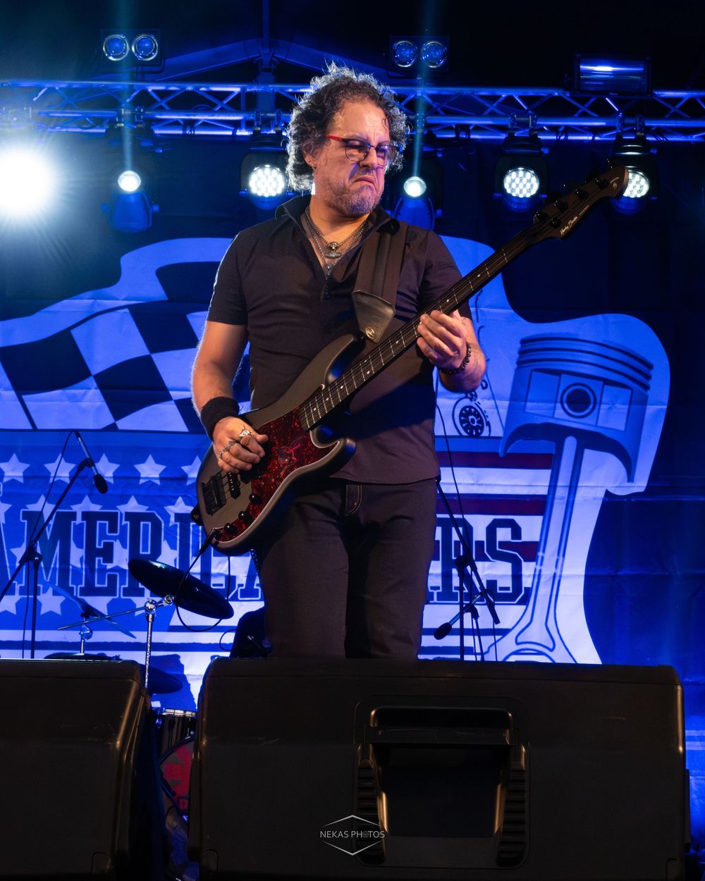 Male guitarist performing on stage with blue lighting and American Riders backdrop at an outdoor concert event.