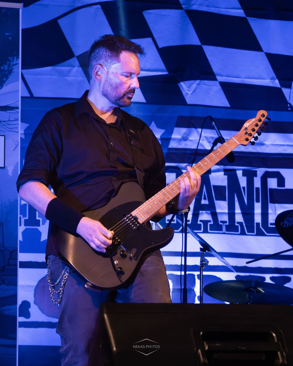 Man in black shirt playing electric guitar on stage with racing flag backdrop under blue stage lighting.