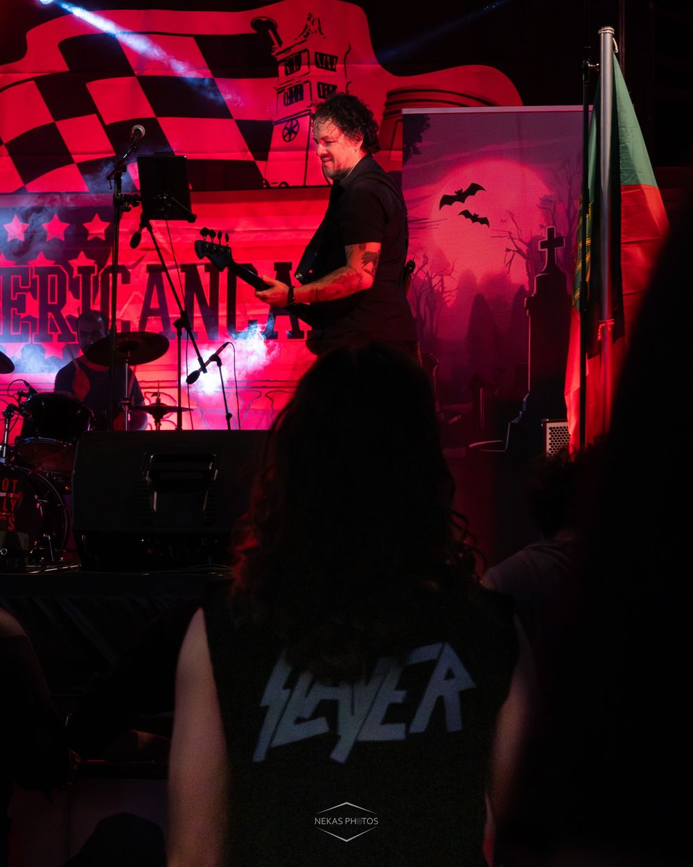 A guitarist performs on a red-lit stage while a fan in a Slayer vest watches from the crowd.