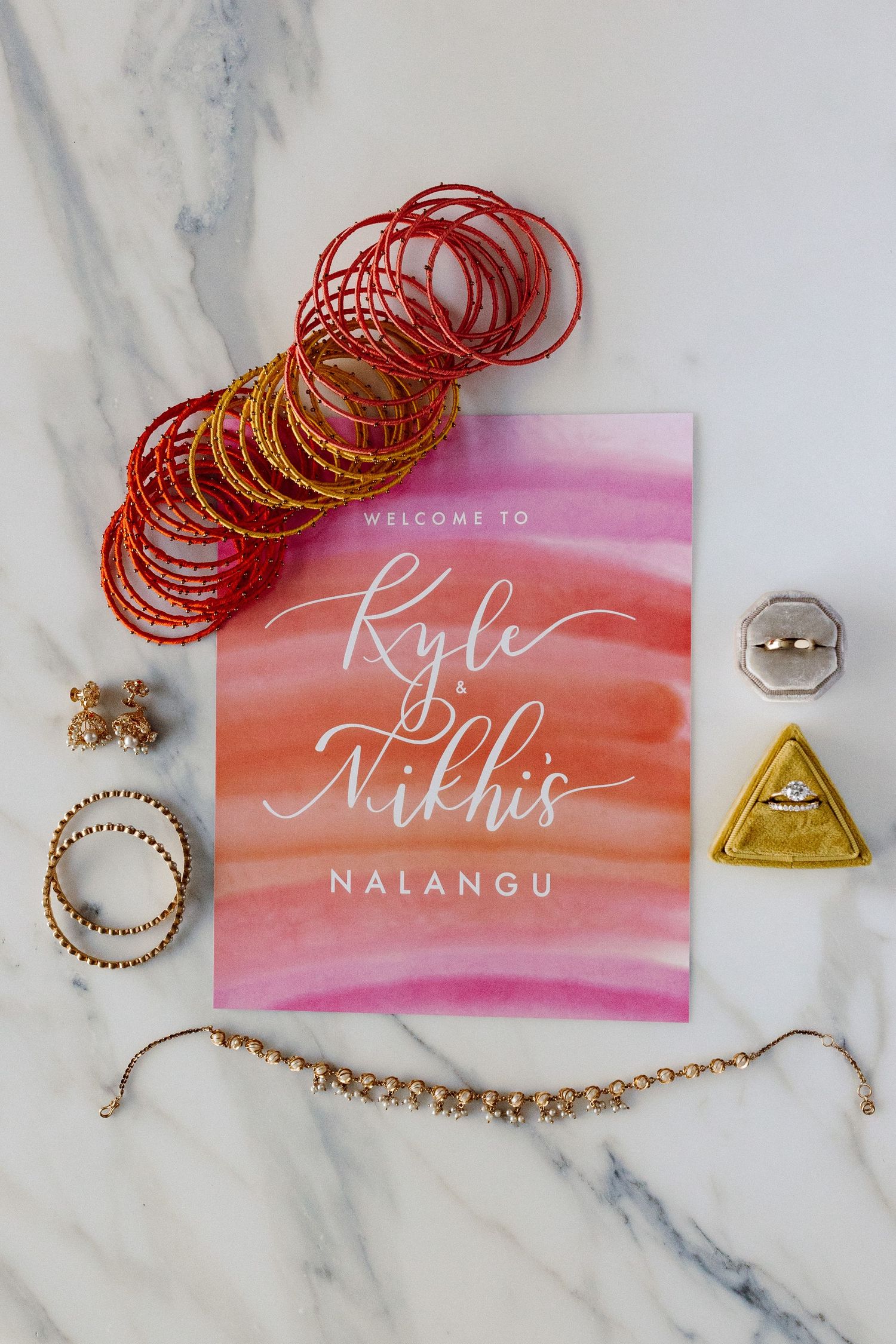 Flat lay of Indian wedding Nalangu invitation with red gold bangles, rings, and jewelry on marble.