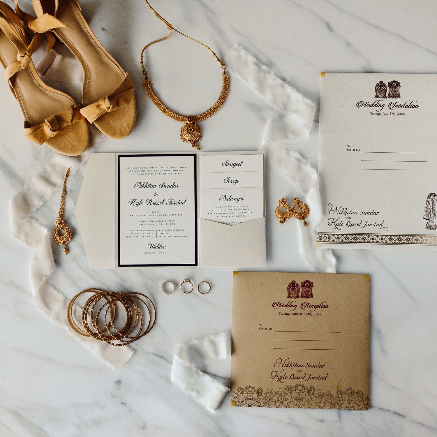 Elegant Indian wedding stationery flat lay with gold jewelry, bangles, rings, and embossed wedding invitations.