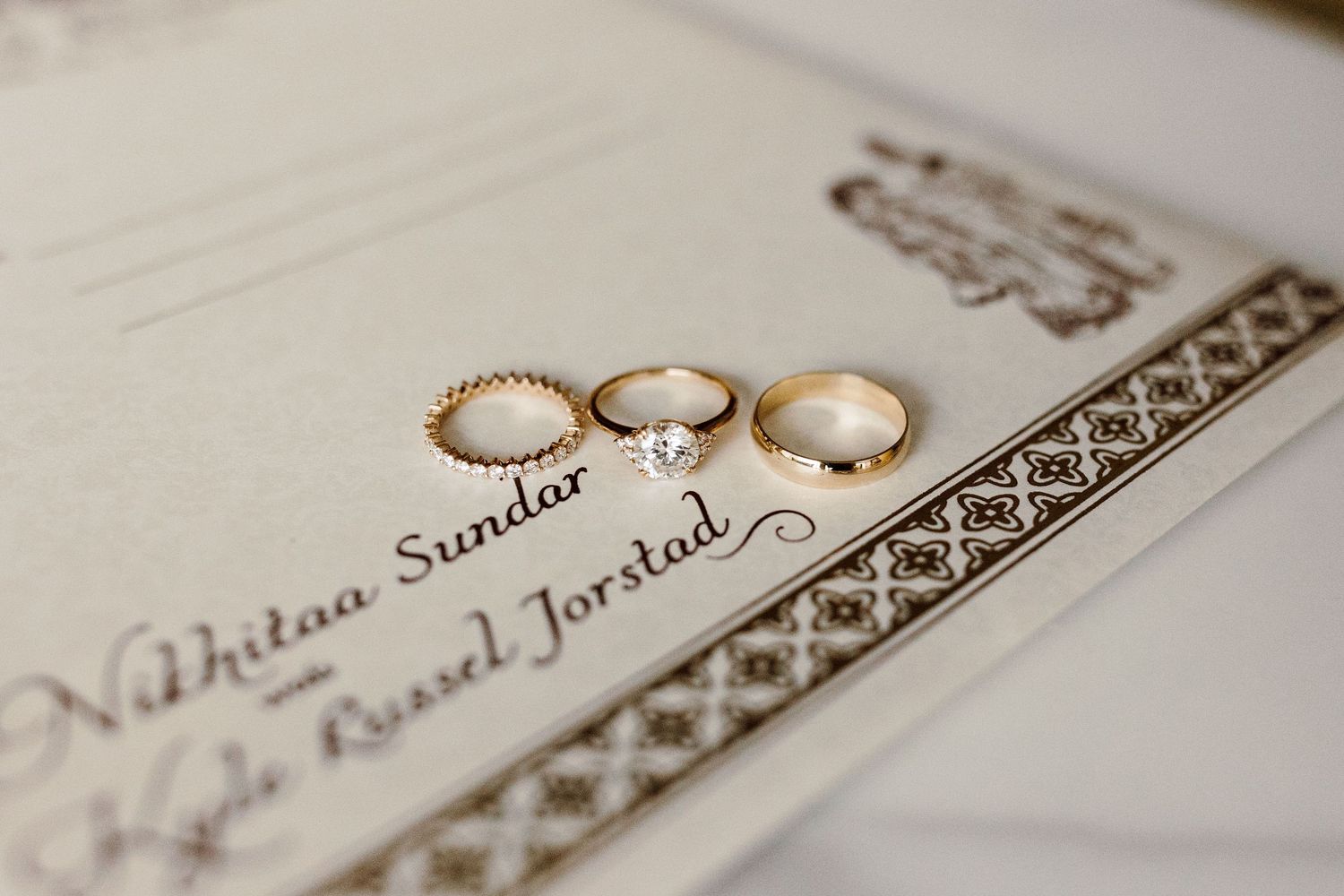 Gold wedding rings and diamond engagement ring displayed on elegant wedding invitation for Nikhitaa Sundar and Russel Jorstad.