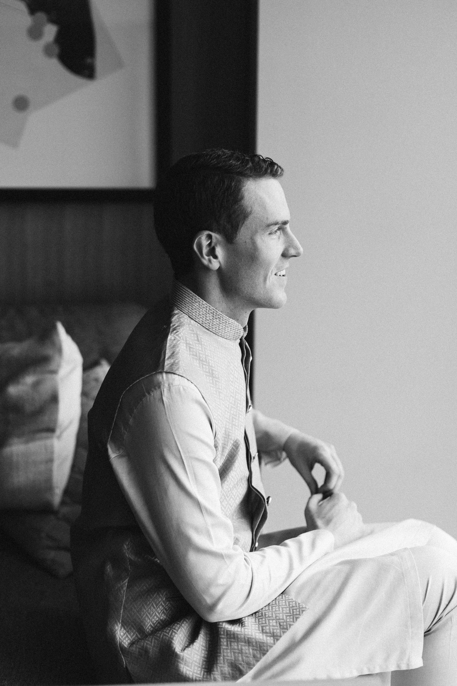 Black and white portrait of a smiling groom in a dress shirt sitting by a window before his wedding.