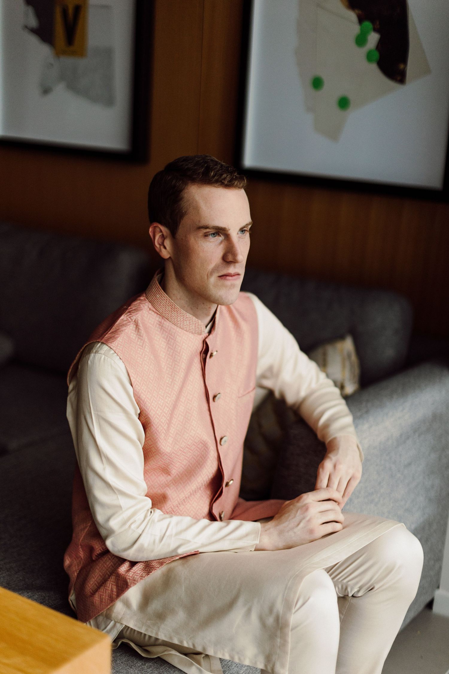 Young man in traditional Indian pink Nehru jacket kurta sitting thoughtfully on a grey sofa.