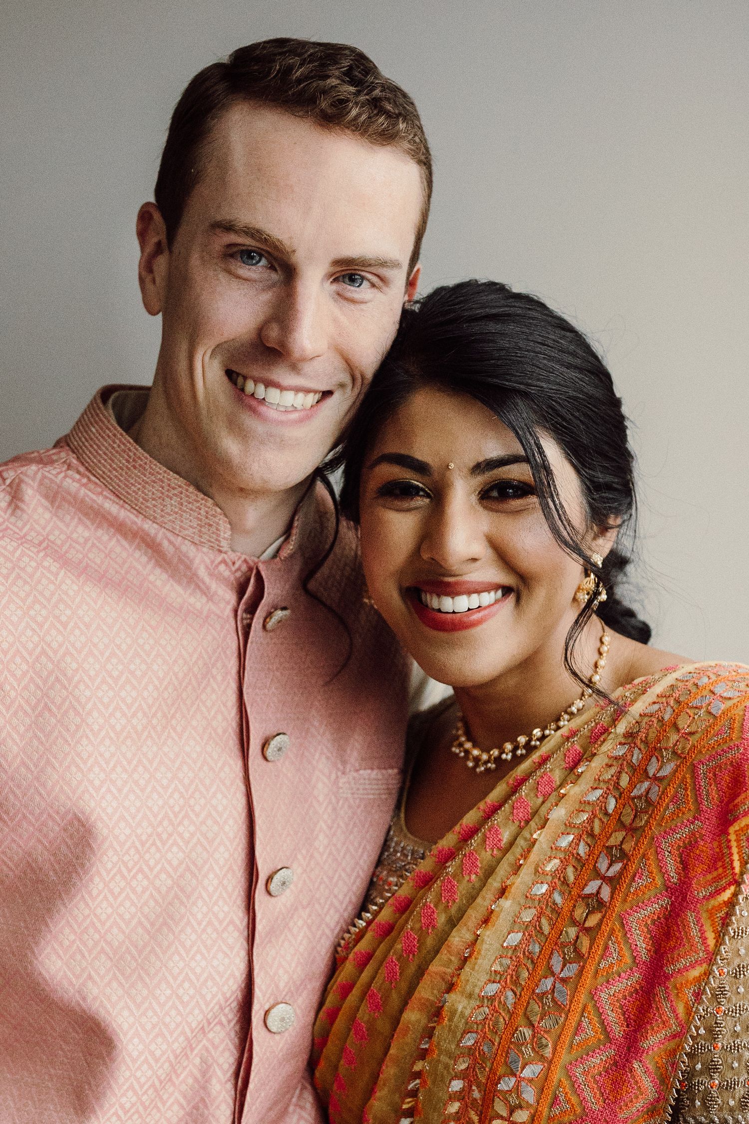 Smiling couple in Indian wedding attire, groom in pink kurta and bride in colorful saree.