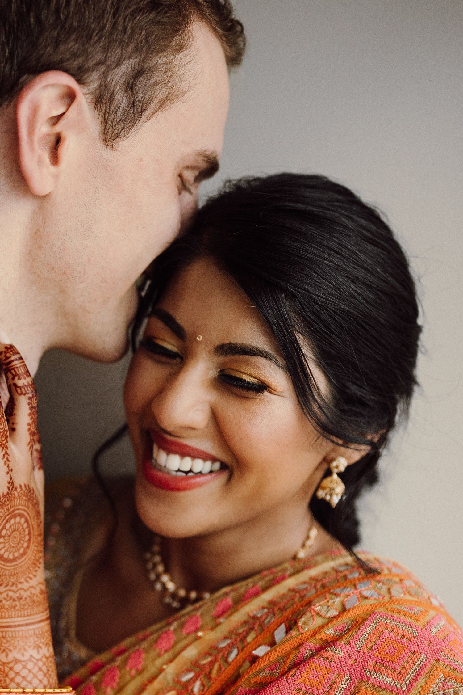 Smiling Indian bride in pink saree with mehndi embraced by groom at multicultural wedding ceremony.